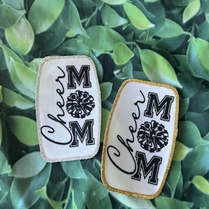 Puede incluir: Dos parches de fieltro blanco con letras negras y un gráfico de animadora en blanco y negro. Los parches dicen "Cheer Mom" y tienen ribetes plateados y dorados.