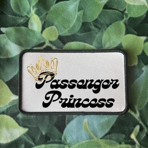 Pode incluir: Patch branco com borda preta e coroa dourada. O texto "Passenger Princess" está em preto com uma fonte retrô.