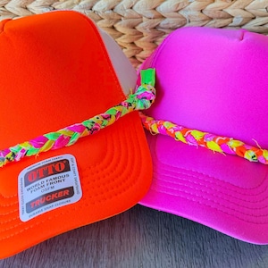 May include: Two trucker hats, one bright orange and one hot pink, with white mesh backs and colorful braided bands. The orange hat has a white tag with black text. The hats are displayed on a wooden surface.