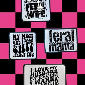 May include: Four white patches with black text on a pink and black checkered background. The patches have phrases like "Somebody's Feral Wife," "My Mom and I Talk Shit About You," "Feral Mama," and "I Love My Husband But Sometimes I Wanna Square Up."