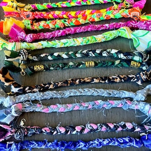 May include: A selection of colourful, braided fabric necklaces. The necklaces display a variety of patterns and colours, including bright pink, green, blue, and patterned fabrics. Each necklace has a small gold-coloured clasp.