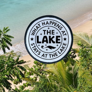 May include: White round patch with black text that reads "What Happens At The Lake Stays At The Lake". The patch has a black border and is on a sandy beach with palm trees.
