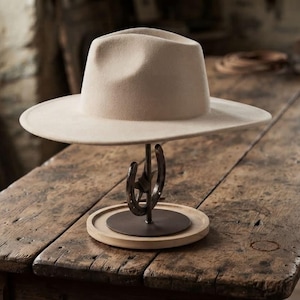May include: A beige felt fedora hat resting on a metal display stand. The stand features a dark metal horseshoe base attached to a circular wooden platform. The hat has a structured crown with a central crease and a wide, flat brim.