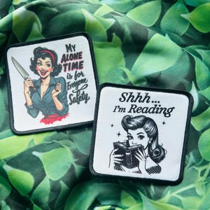May include: Two iron-on patches with black borders. The first patch features a cartoon woman with a knife and the text "My Alone Time is for Everyone Safety". The second patch features a cartoon woman reading a book with the text "Shhh... I'm Reading".