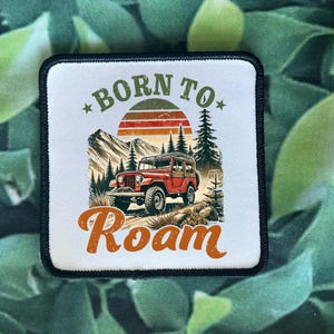 May include: Square patch with a black border featuring a red Jeep driving through a mountain landscape. The patch has the text "BORN TO" above a sunset graphic and "Roam" below.