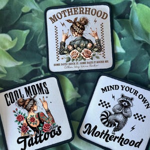 May include: Three iron-on patches with white backgrounds and black borders. The top patch features a woman with a bandana and the text "Motherhood. Some days I rock it. Some days it rocks me. Either way we're rockin". The left patch features a woman with tattoos and the text "Cool Moms Have Tattoos". The right patch features a raccoon with the text "Mind Your Own Motherhood".