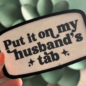 May include: White fabric patch with black text that reads "Put it on my husband's tab" with two small black diamond shapes on either side of the text.