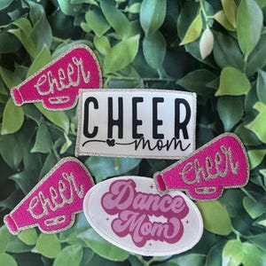 Peut inclure: Quatre patchs thermocollants sur le thème du cheerleading. Un patch dit "Cheer Mom" en lettres blanches avec un contour argenté pailleté. Un autre patch dit "Dance Mom" en lettres roses avec un contour blanc. Les deux autres patchs sont des mégaphones roses avec un contour argenté pailleté et le mot "Cheer" écrit dessus.