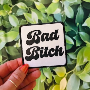 May include: A white square patch with black embroidered text that reads "Bad Bitch" in a retro font style.