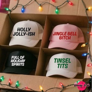 May include: Four trucker hats in a cardboard box. The top left is white with black text reading HOLLY JOLLY-ISH. The top right is pink with red text reading JINGLE BELL BITCH. The bottom left is black with white text reading FULL OF HOLIDAY SPIRITS. The bottom right is white with green text reading TINSEL TITS.