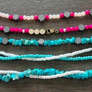 May include: A selection of beaded necklaces in different designs. One necklace spells "OUTLAW" in gold letter beads. Other necklaces feature pink, white, and turquoise beads, with checkered pattern accents.