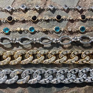 May include: An assortment of necklaces and bracelets with various designs. The jewelry includes chains with clear rhinestones, black and teal colored stones, and gold and silver tones. The pieces are displayed on a textured, dark surface.