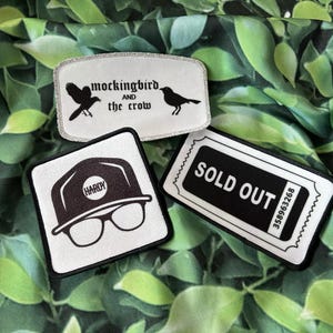 May include: Three embroidered patches with black and white designs. One patch reads "mockingbird and the crow" with bird illustrations. Another features a hat and glasses with the name "Hardy." The third says "SOLD OUT."