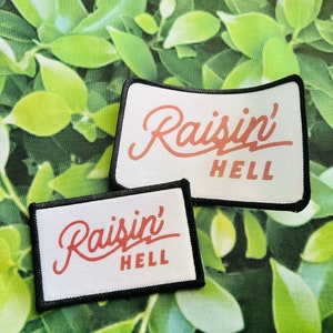 May include: Two white fabric patches with black stitching. Both patches have the text "Raisin' Hell" in red cursive lettering.