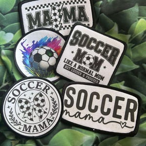 May include: Four iron-on patches with a soccer theme. The patches are white with black text and designs. The text on the patches reads "Mama", "Soccer Mom", "Soccer", and "Soccer Mama".