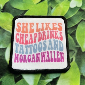 May include: A white patch with black stitching around the edges. The patch has a colorful retro font design that reads "She Likes Cheap Drinks Tattoos and Morgan Wallen".