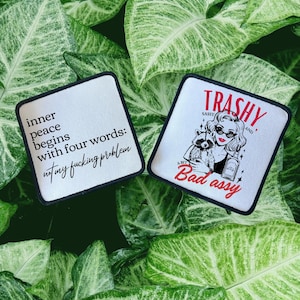 May include: Two white iron-on patches with black text and images. The first patch says "inner peace begins with four words: not my fucking problem". The second patch says "Trashy and a bit bad assy" with a cartoon image of a woman with a raccoon.