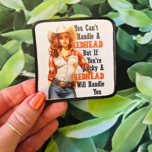 May include: Square patch with a pin-up style illustration of a woman in a cowboy hat and plaid shirt. The text reads: "You Can't Handle A REDHEAD But If You're Lucky A REDHEAD Will Handle You."