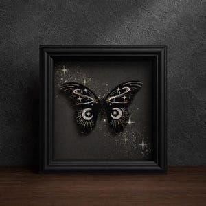 May include: A black butterfly specimen displayed in a square black frame. The butterfly wings feature intricate white and gold celestial patterns, including crescent moons, stars, and orbital rings, set against a dark background with star dust.