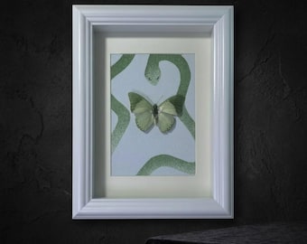 May include: A framed artwork featuring a pale green butterfly centred on a light blue background with a dark green snake pattern. The piece is enclosed in a white, multi-layered rectangular frame with a clean, structured border design.
