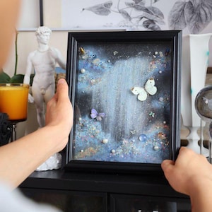 May include: A framed art piece featuring a waterfall scene with butterflies and decorative elements. The artwork has a black frame and a dark blue and white color scheme. The butterflies are white and purple.