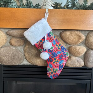 Peut inclure: Une chaussette de Noël rouge et bleue à fleurs avec des pompons blancs suspendue à une cheminée.