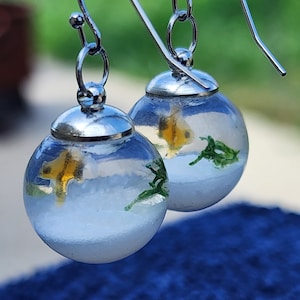 May include: A pair of silver earrings with clear glass globes containing a small yellow fish and green plant. The globes are suspended from silver hooks.