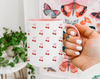 May include: A pink and white ceramic mug with a pattern of pink cherries. The mug has a pink rim and handle.