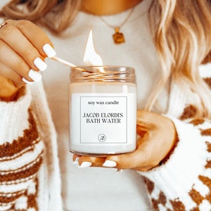 May include: A white soy wax candle with a label that reads "soy wax candle" and "JACOB ELORDI'S BATH WATER".