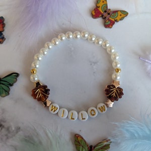 Könnte beinhalten: Ein weißes Perlenarmband mit goldenen Akzenten und braunen Blattanhängern. Das Armband buchstabiert den Namen "WILLOW" in weißen Buchstaben mit goldenen Akzenten.