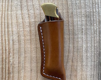 May include: A folding pocket knife with a brass bolster and wood handle is partially inserted into a smooth, tan leather sheath. The sheath features visible white stitching along the side and bottom edge, securing the knife in a vertical position.