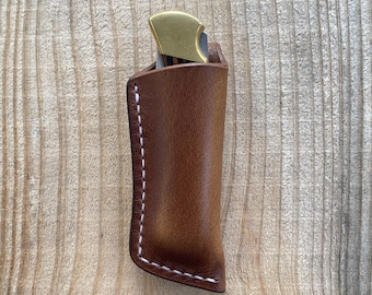 May include: A brown leather sheath containing a folding knife with a brass bolster. The sheath features white stitching along the side and bottom edges, securely holding the knife handle which is partially visible at the top of the leather pouch.