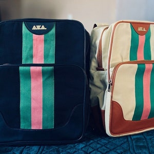 May include: Two canvas backpacks with green, pink, and brown stripes. The backpacks have the letters "AKA" in gold on the front.