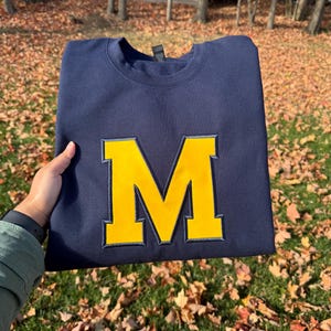 Peut inclure: Sweat-shirt bleu marine avec un grand logo "M" jaune. Le "M" est bordé de bleu marine. Le sweat-shirt est présenté sur un fond de feuilles d'automne et d'herbe verte.