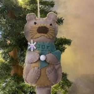 May include: A brown felt ornament shaped like an otter wearing a green scarf with the word "JOY" on it. The otter is holding a white snowflake.