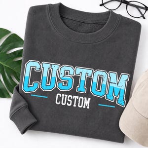 Può includere: Una felpa girocollo grigio antracite scuro con la scritta CUSTOM in grandi lettere stampatello azzurre dall'aspetto usurato con un contorno bianco, posizionata sopra una versione più piccola del testo CUSTOM in bianco con linee orizzontali.