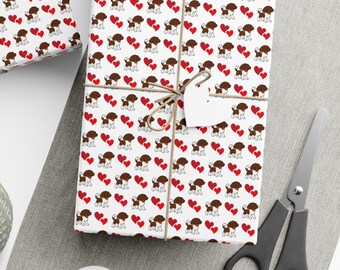 Puede incluir: Papel de regalo blanco con un patrón repetido de perros marrones y corazones rojos. El regalo está atado con hilo y una etiqueta en forma de corazón. También se ven unas tijeras y etiquetas en blanco.