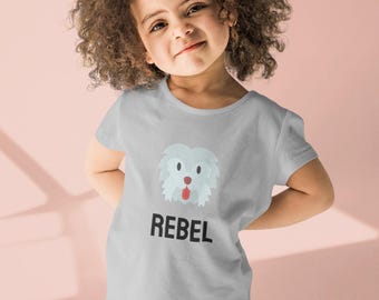 May include: A light gray t-shirt featuring a cartoon dog face with the word "REBEL" in black block letters. The shirt is worn by a young child with curly hair and blue jeans.