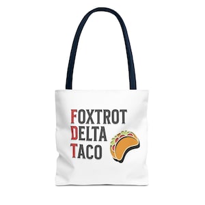 May include: White tote bag with navy blue handles featuring the text "Foxtrot Delta Taco" in red and black, with a cartoon taco graphic. The bag is designed for carrying items.
