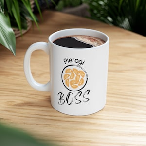 May include: White ceramic mug with a black handle, filled with dark coffee. The mug features the text "Pierogi BOSS" with an illustration of pierogi. The mug is on a wooden surface.