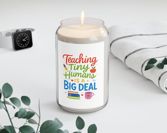 May include: A lit candle in a clear glass jar with a white label. The label reads "Teaching Tiny Humans is a BIG DEAL" in colorful text. A white watch and a folded white and gray striped towel are in the background.
