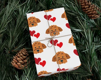 Puede incluir: Papel de regalo blanco con un patrón repetido de caras de perro marrón y corazones rojos. El paquete envuelto está atado con hilo y descansa sobre agujas de pino y piñas verdes.