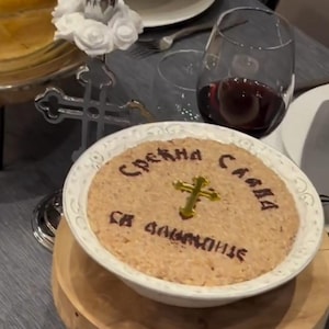 Peut inclure: Un bol en céramique blanche rempli d'une garniture brune et surmonté d'une croix dorée. Le bol est posé sur un support en bois avec des pieds en métal noir. Le bol porte l'inscription "Срећна слава са благословије" écrite en brun sur la garniture.