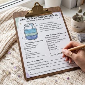 May include: A wooden clipboard with a printed worksheet titled "When My Brain Feels Overwhelmed." The worksheet includes sections on causes and symptoms of overwhelm, and how to cope. A hand is writing on the worksheet with a pen.