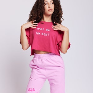 May include: A woman wearing a pink cropped t-shirt with the text "MAKE HIM PAY RENT" and pink sweatpants with the text "ALL THINGS BAD" in a retro font.