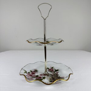 May include: Two-tiered serving tray with a silver handle and stem. The clear glass plates have a scalloped edge with gold trim and are decorated with pink floral patterns. A silver chain rests on the bottom tier.