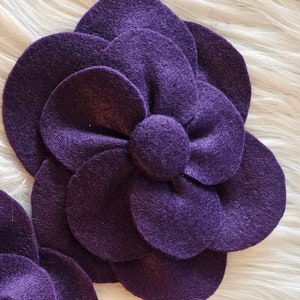 May include: A large purple felt flower with multiple layers and a center bow. The flower is made of felt and has a soft, fuzzy texture.