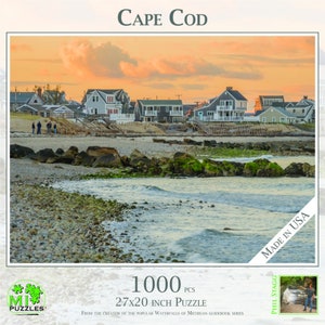 May include: A 1000 piece jigsaw puzzle featuring a scenic sunset view of Cape Cod, Massachusetts. The puzzle depicts a row of houses along a sandy beach with rocky outcroppings and calm water. The puzzle is made in the USA and measures 27x20 inches.