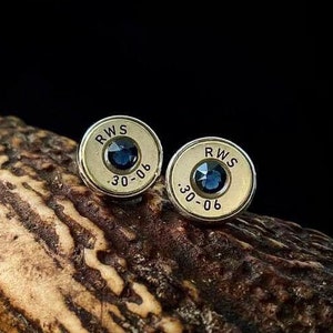 Peut inclure: Une paire de boucles d'oreilles en forme de douille de balle dorée avec des pierres précieuses bleues et le texte "RWS 30-06" gravé sur chaque boucle d'oreille.