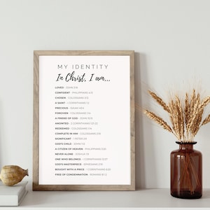 May include: A framed print with the text "MY IDENTITY In Christ, I am..." and a list of affirmations. The frame is light brown. A brown glass vase with wheat stalks is to the right. A small, decorative object sits on a white surface.